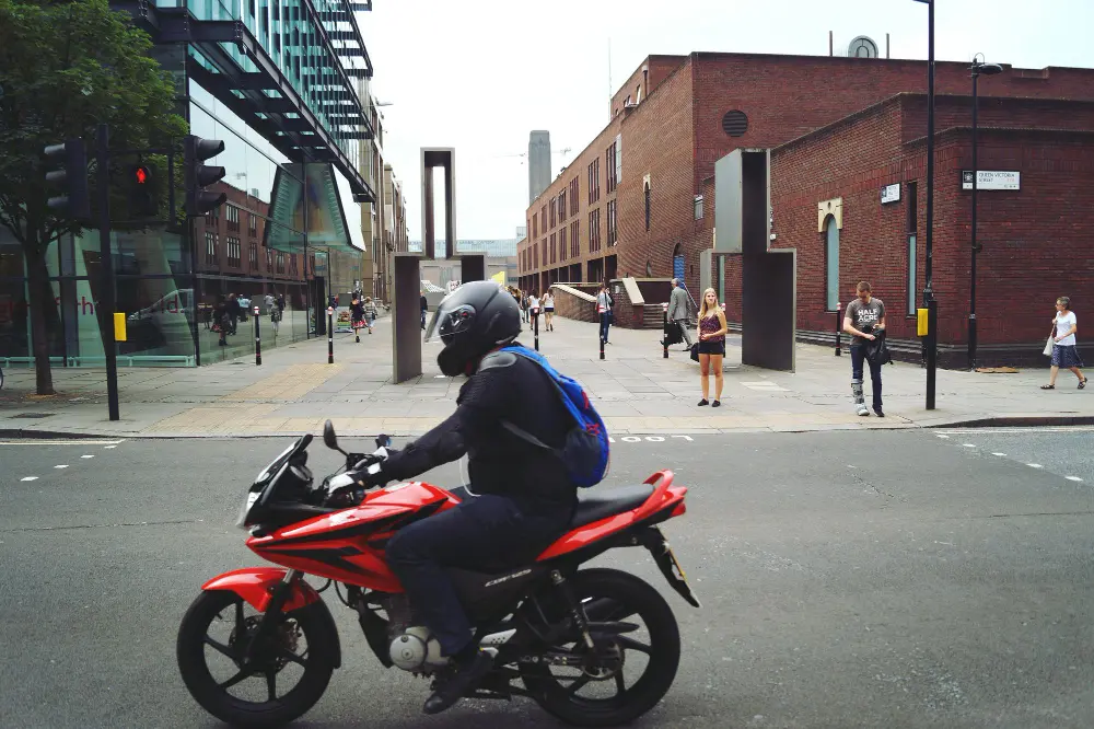 Homem pilotando uma moto. Ele veste roupas de inverno, capacete e tem uma mochila nas costas. Ele passa por uma região que parece urbana.