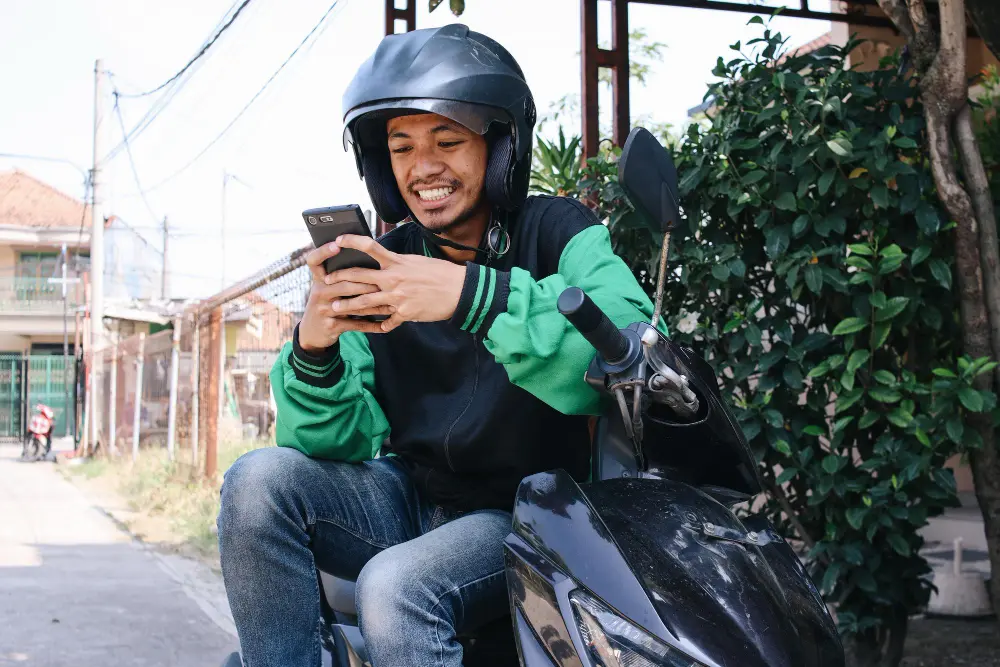 Homem sentado em uma motocicleta olhando para seu smartphone, que segura com as mãos. O homem sorri olhando para a tela, demonstrando felicidade. Ele veste calça jeans, casaco e está de capacete. Ao fundo, vê-se uma rua.