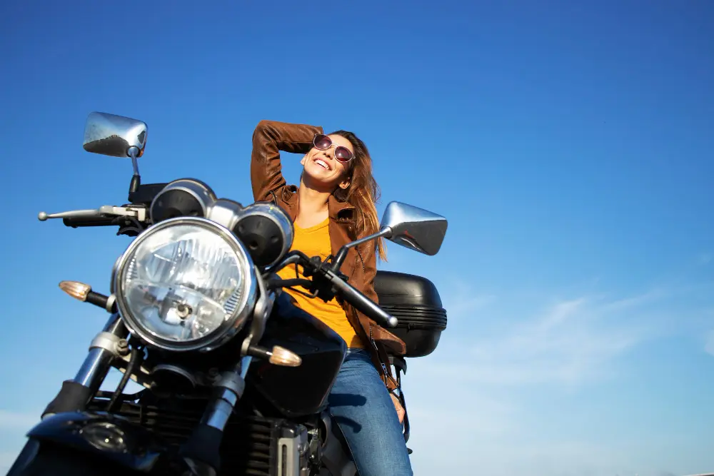 Mulher sentada em uma moto. Ela está feliz, sorrindo, usando calça jeans, blusa amarela e casaco marrom. Ela também usa um óculos de sol. Ao fundo vemos o céu azul.