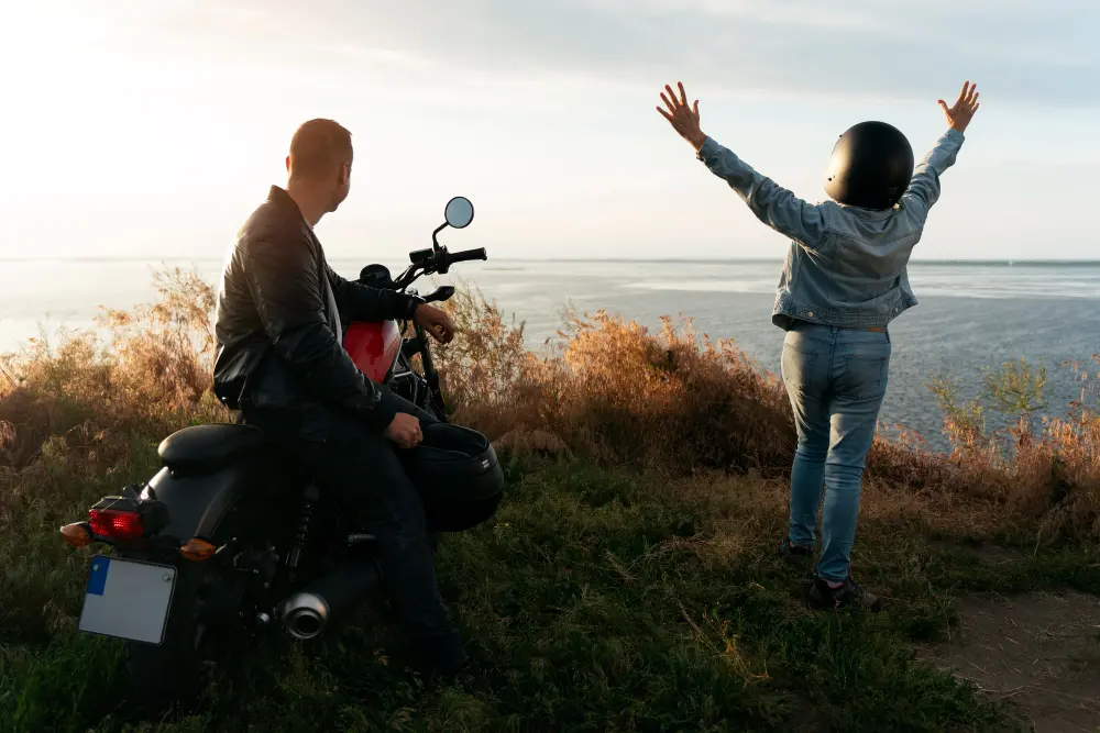 Duas pessoas de frente para uma paisagem. O homem está sentado na moto, olhando o horizonte. A mulher está de pé, com os braços abertos e usando capacete, demonstrando felicidade. A paisagem é um monte e ao fundo há o mar.