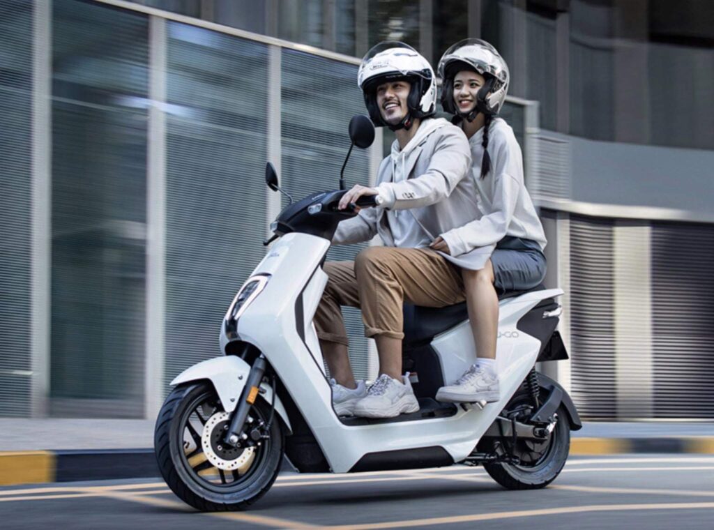 Casal andando em uma moto elétrica branca. Eles estão usando capacetes e parecem estar felizes.
